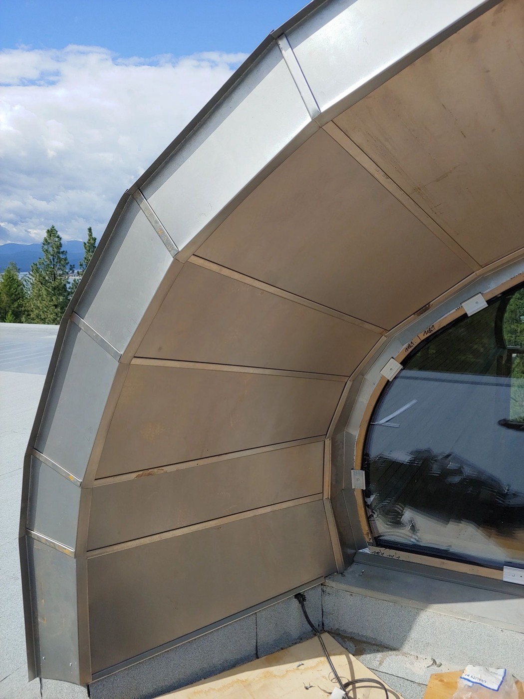 Close-up of curved standing-seam metal panels wrapping an arched eyebrow dormer, with mountains behind